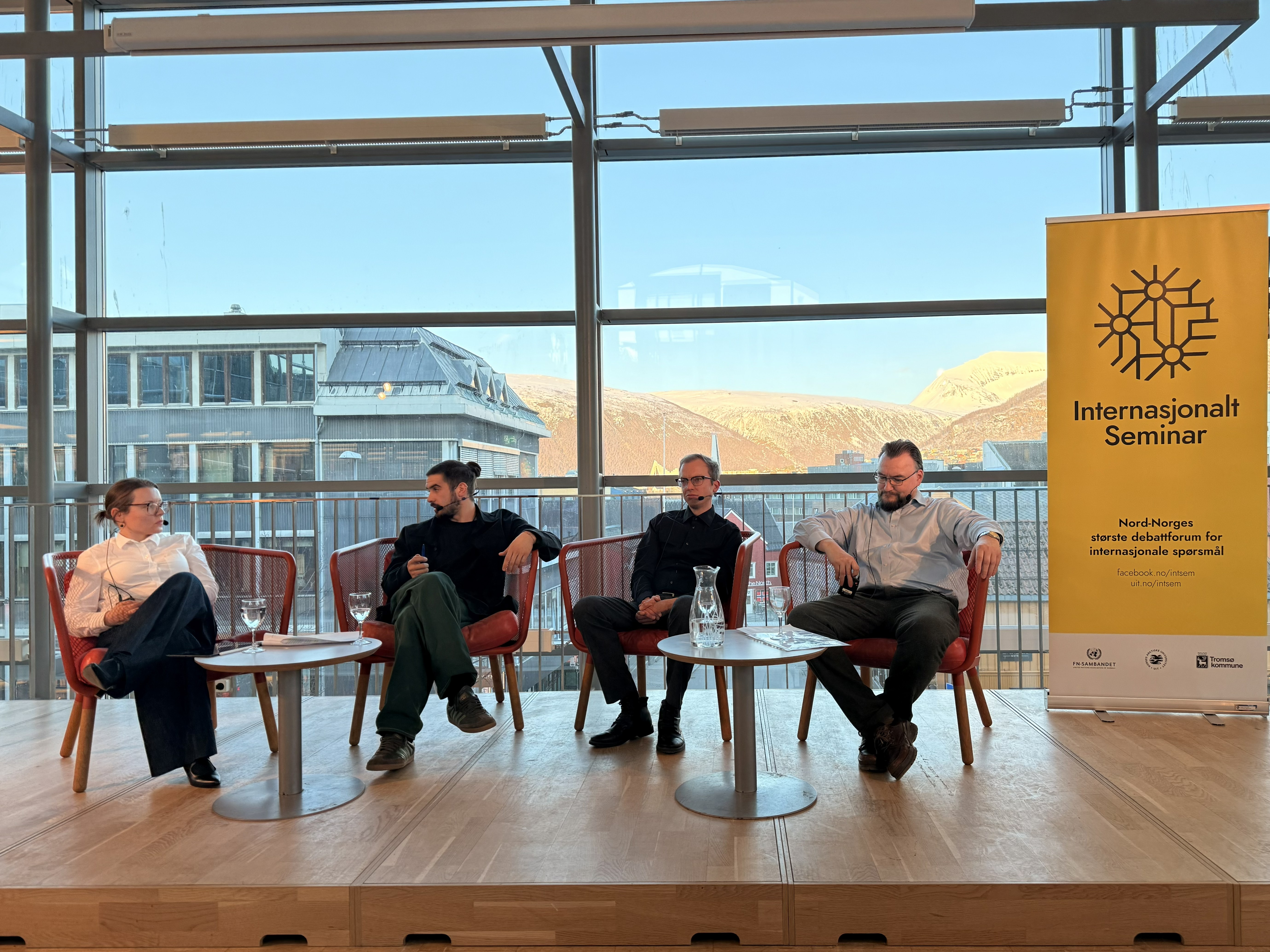 woman, wearing white shirt, sitting on a stage, next to her a man, and next to him, two other menall wearing shirts, sitting in red chairs, large windows with natural lighting in the background